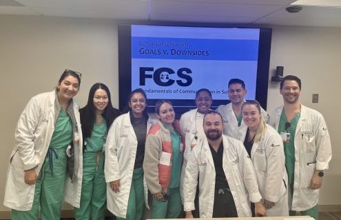 Surgical residents in green scrubs and white coats stand in front of an FCS curriculum slide.